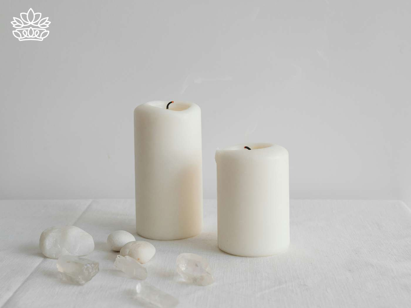 Two unlit ivory pillar candles accompanied by a scattering of translucent crystals on a clean white cloth, a minimalist addition to the Candles Collection - Fabulous Flowers and Gifts.