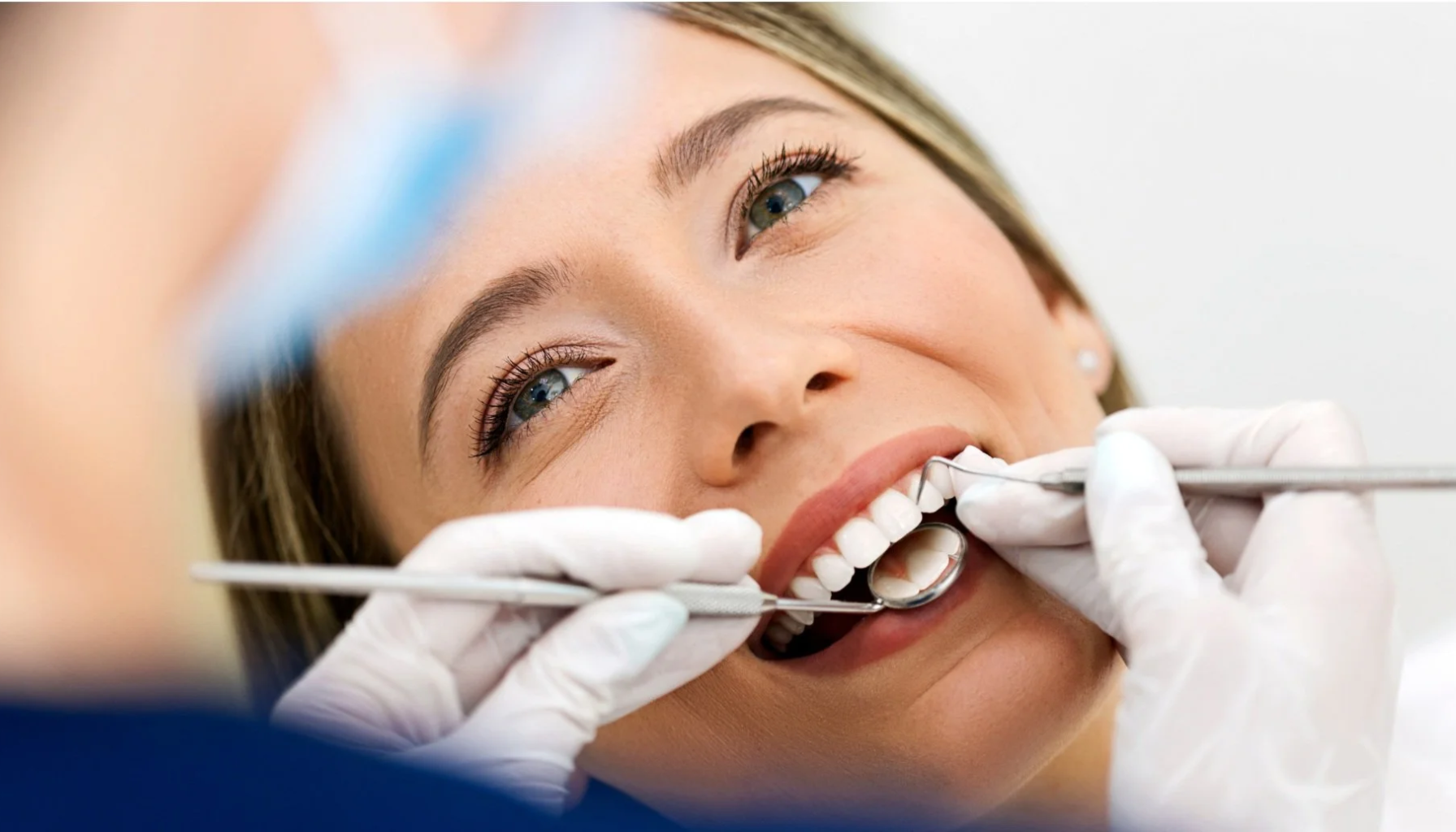 "Kvinne ligger i tannlegestol mens tannlege undersøker tennene hennes med speil og instrument."