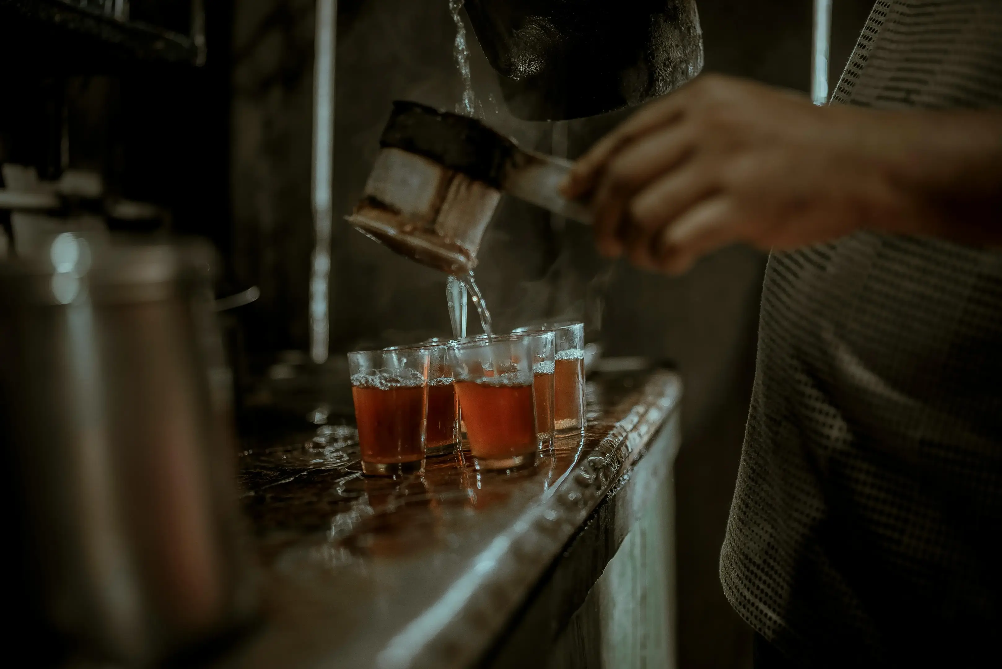 A person pours a dark, steaming liquid through a traditional cloth strainer into several small glasses lined up on a rustic counter. The scene is captured in a moody, low-light setting with a shallow depth of field that highlights the motion of the pour.