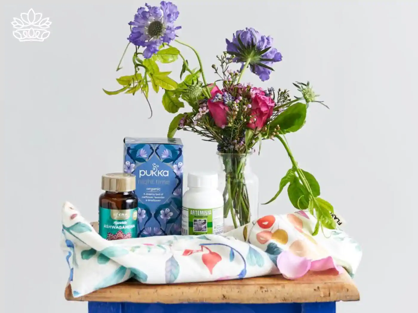 A collection of natural wellness products including Pukka Night Time tea, ashwagandha supplement, and Artemisia absinthium tincture, arranged on a wooden tray with vibrant fresh flowers. Healthy Gift Boxes Delivered with Heart. Fabulous Flowers and Gifts.