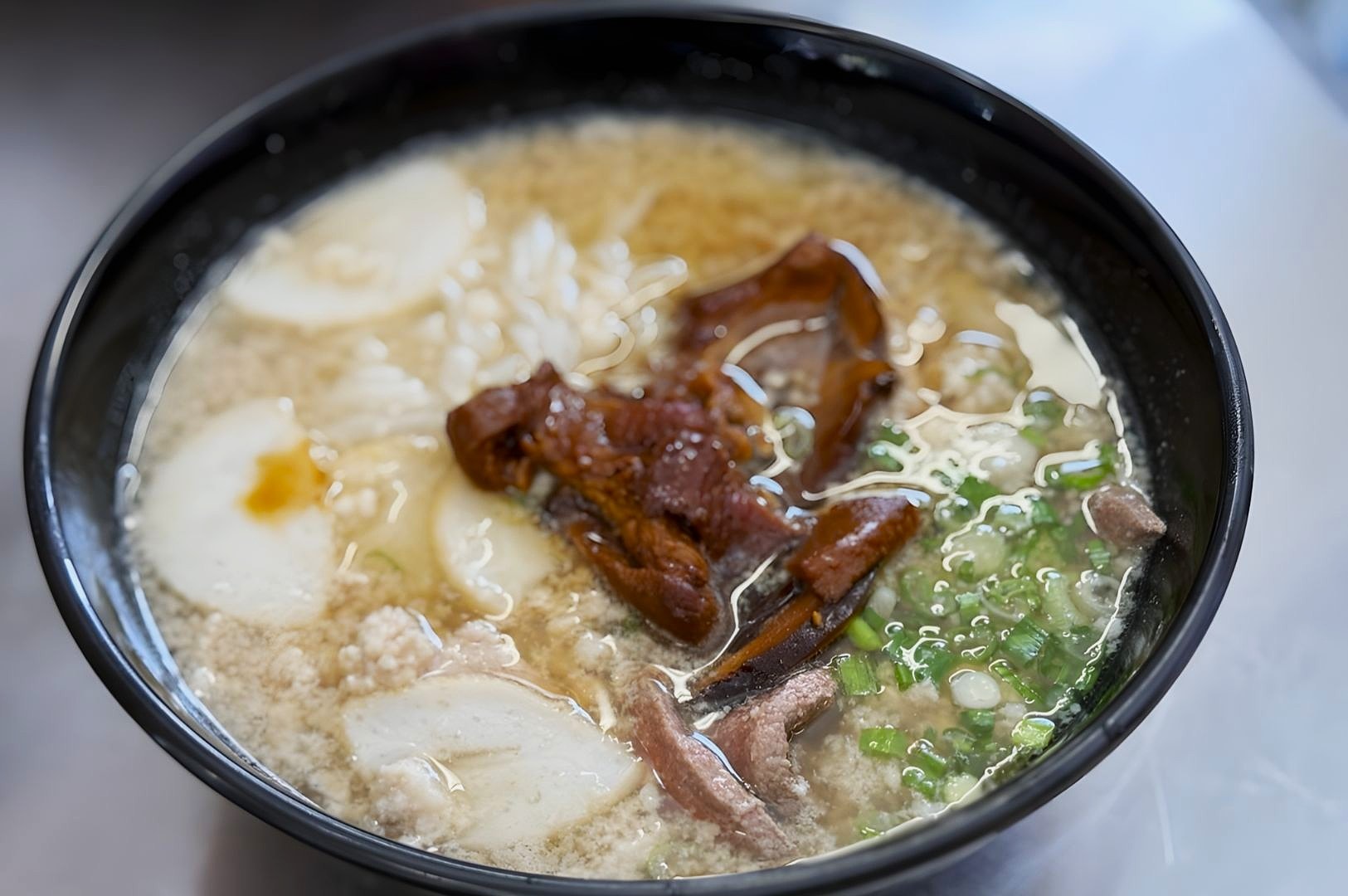A black bowl filled with rich ramen soup features pork slices, green onions, bamboo shoots, and fish cakes, creating a warm and savory appearance.