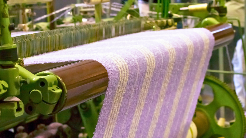 A loom weaving a fabric with light purple and white stripes.