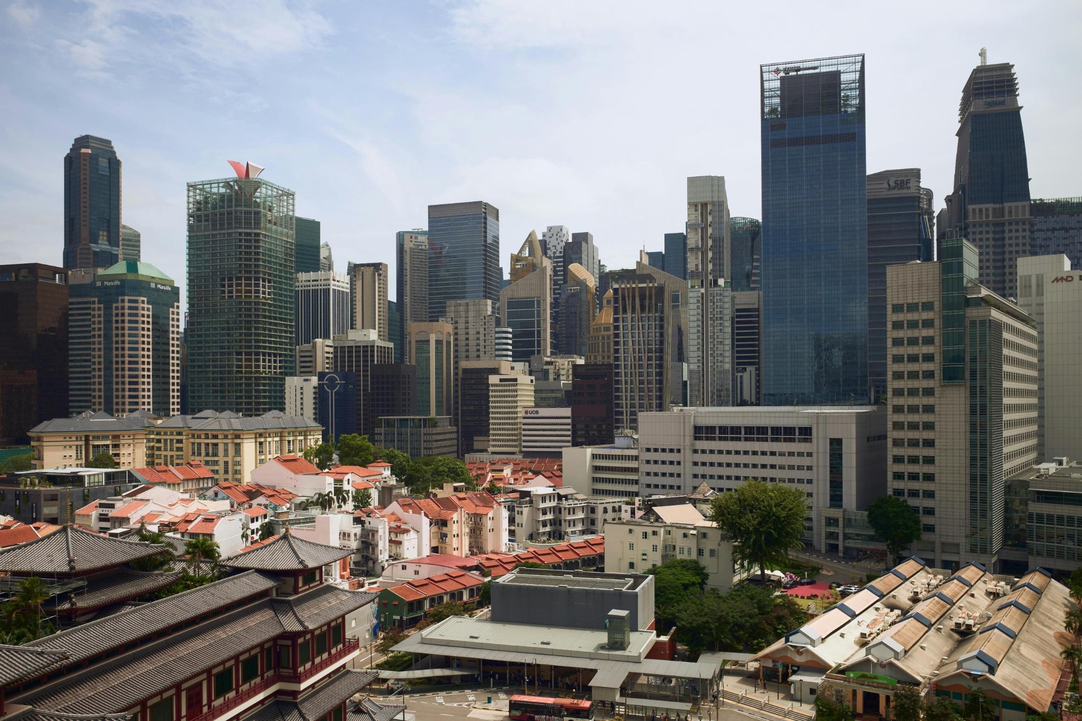 This image features the ornate, tiered blue roof of a Chinese pavilion topped with a golden finial, framed by a tall tree with reddish-brown foliage on the left. To the right, a section of green tiled roofing displays mythical figurines along the ridge, while a faint moon hangs in the clear blue sky between the architectural structures.