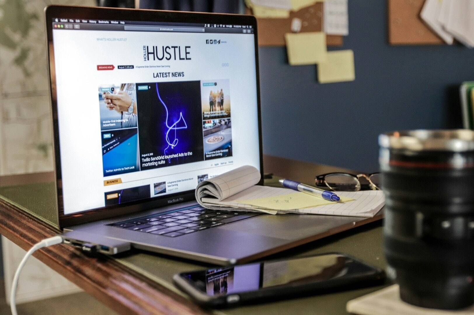 A laptop displaying "The Hustle" news site sits on a cluttered desk with a notepad, pen, eyeglasses, smartphone, and coffee cup, evoking a busy work atmosphere.