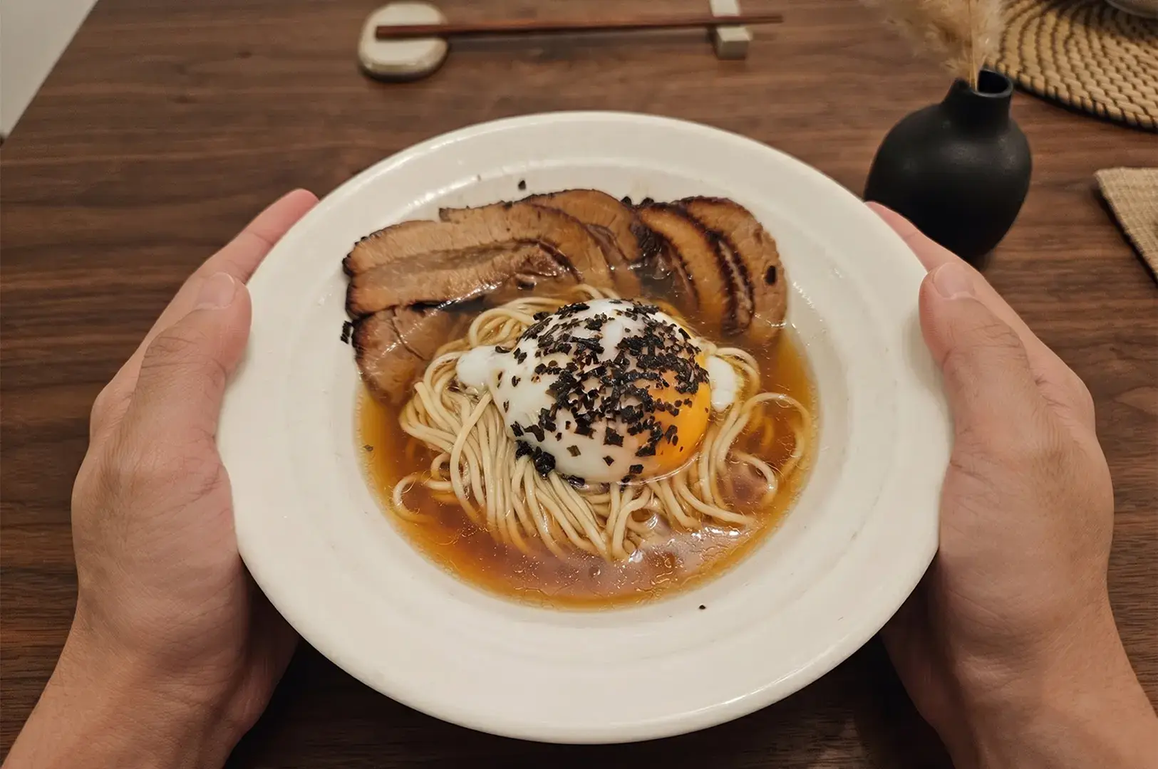 Hands holding a bowl of homemade ramen with chashu pork, a soft-boiled egg, and seaweed topping.