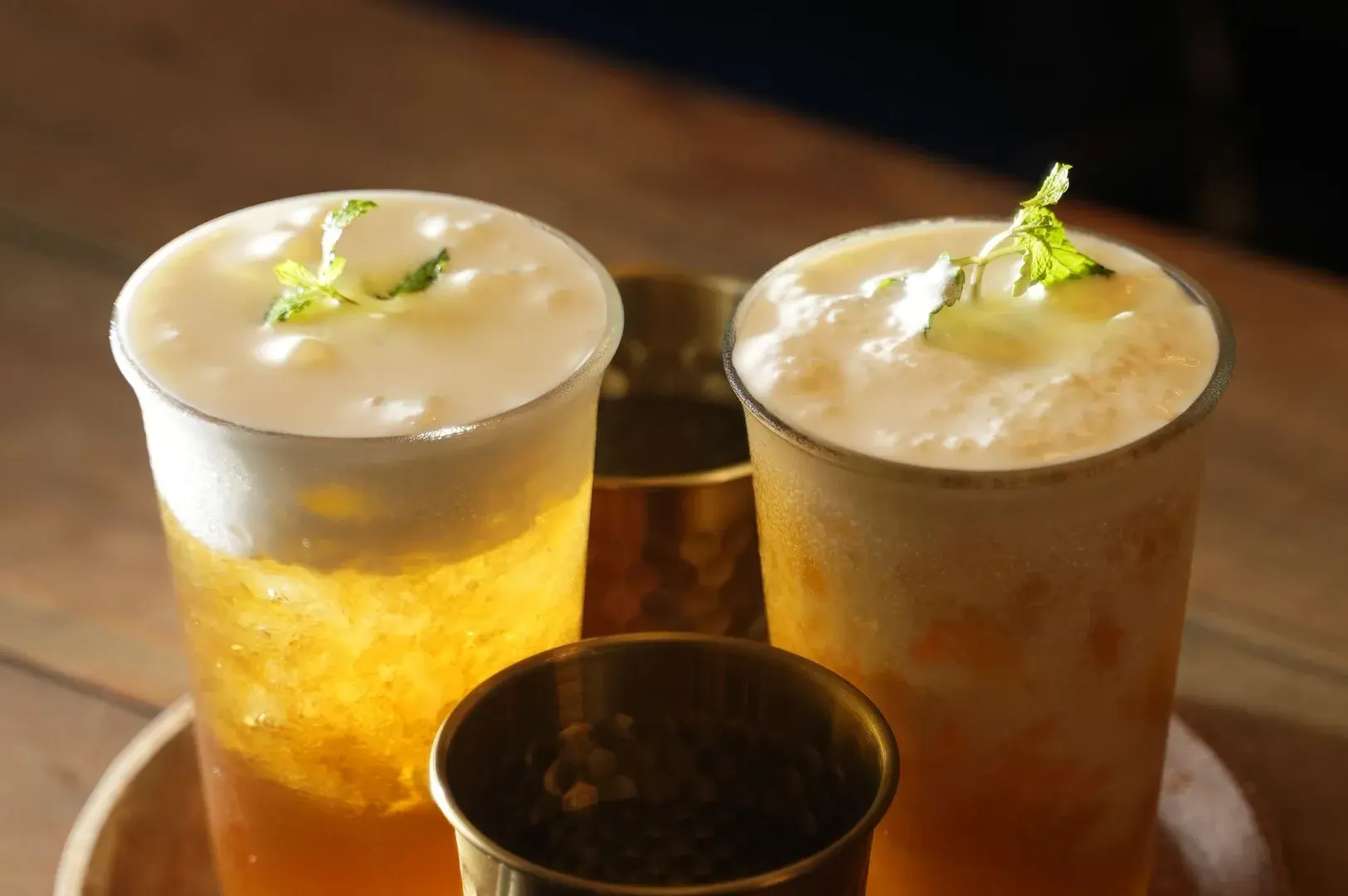 Two tall cups of iced tea topped with thick milk foam and a mint sprig, served on a tray with small containers.