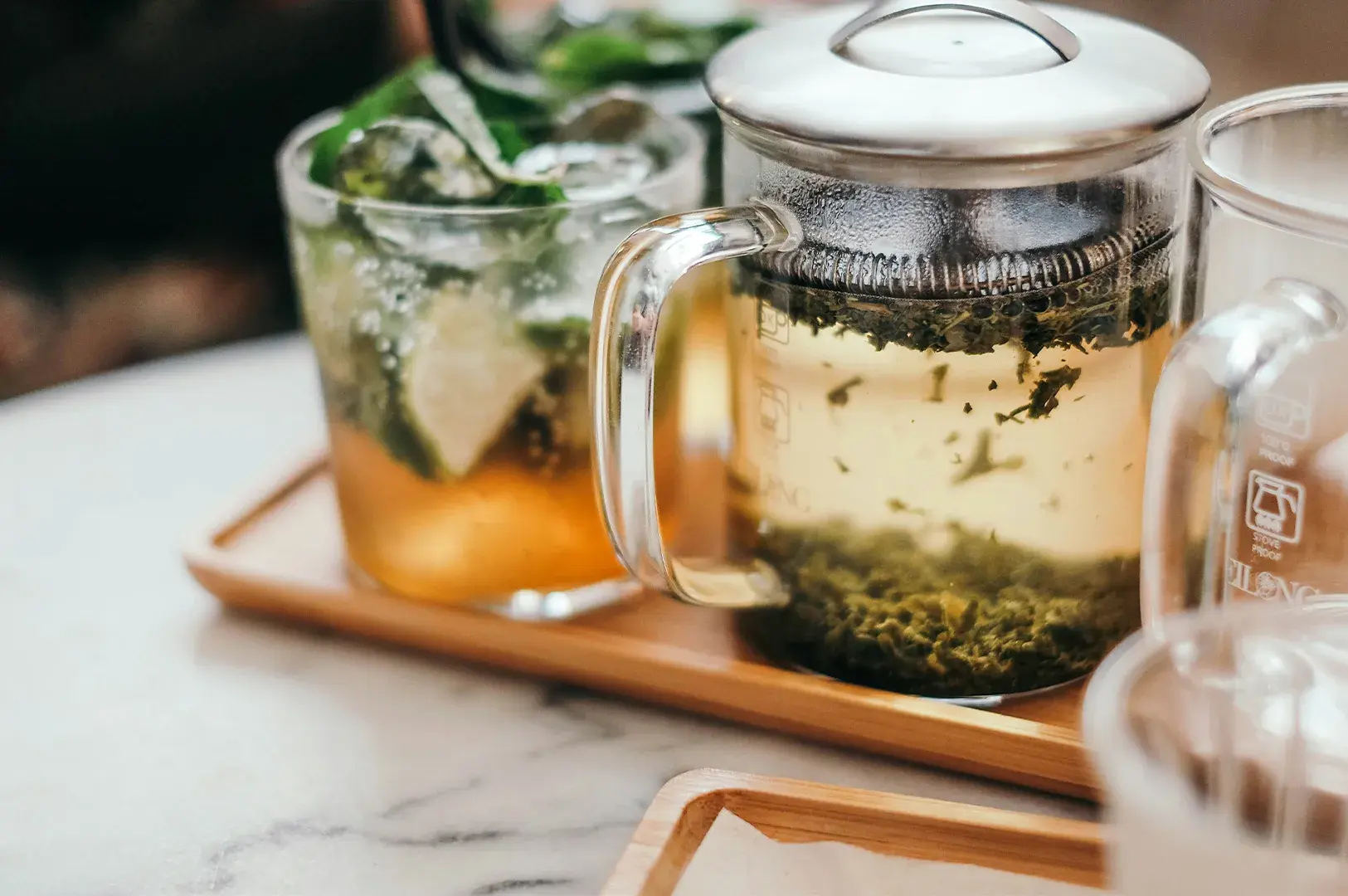 Glass teapot with loose-leaf green tea and iced mint beverage on a wooden tray.