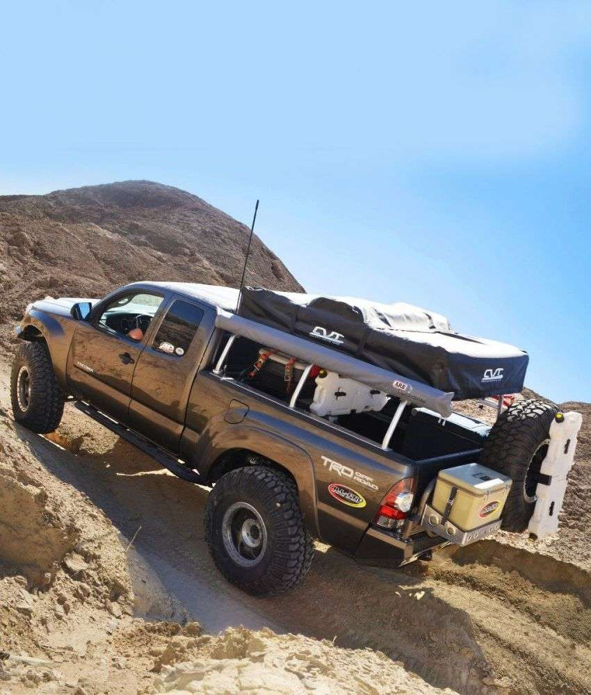 All-Pro Off-Road Bed Rack with tent and spare tire on Toyota Tacoma driving up a dirt road.