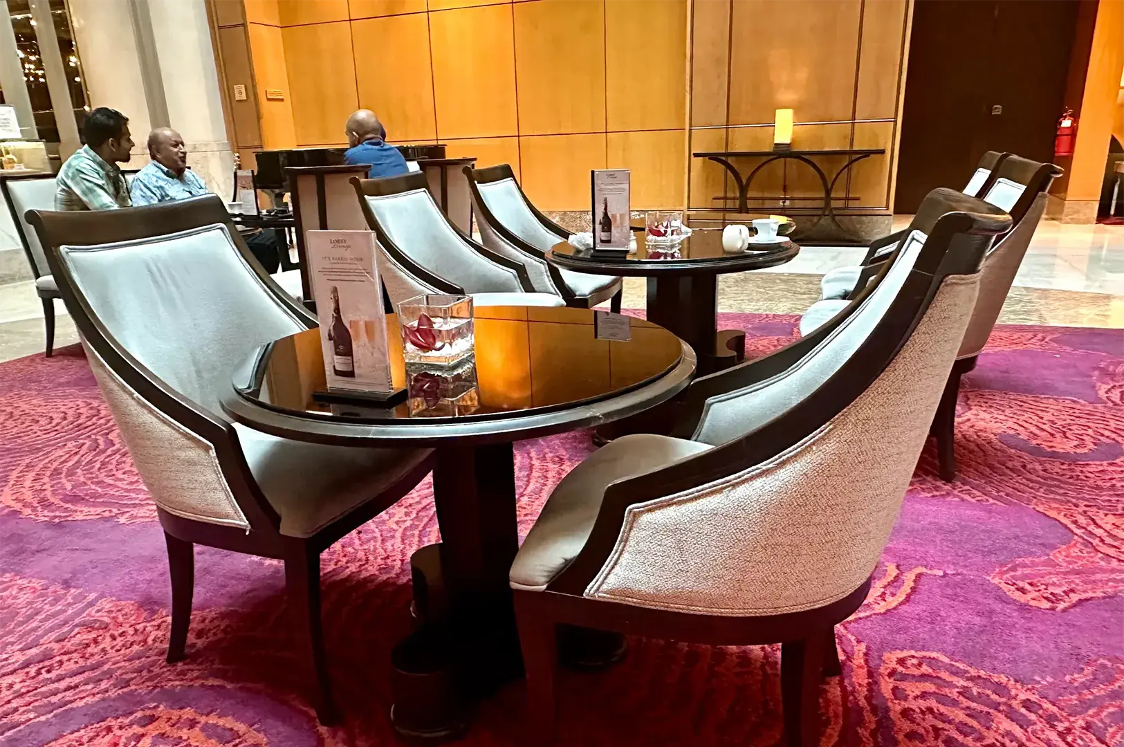 An elegant hotel lobby lounge area featuring comfortable upholstered armchairs, circular glass-topped wooden tables, and a vibrant purple pattern carpet.