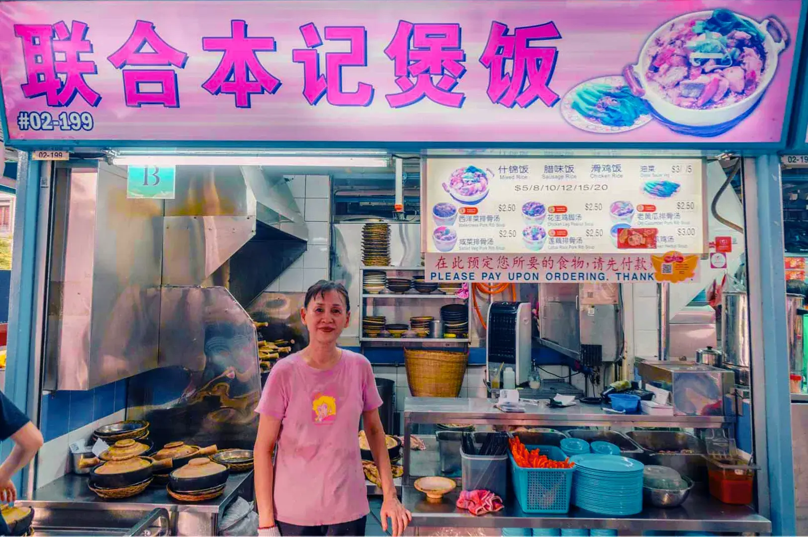 A smiling vendor stands in front of a brightly colored food stall, adorned with a pink sign featuring Chinese characters. Various dishes and prices are displayed.