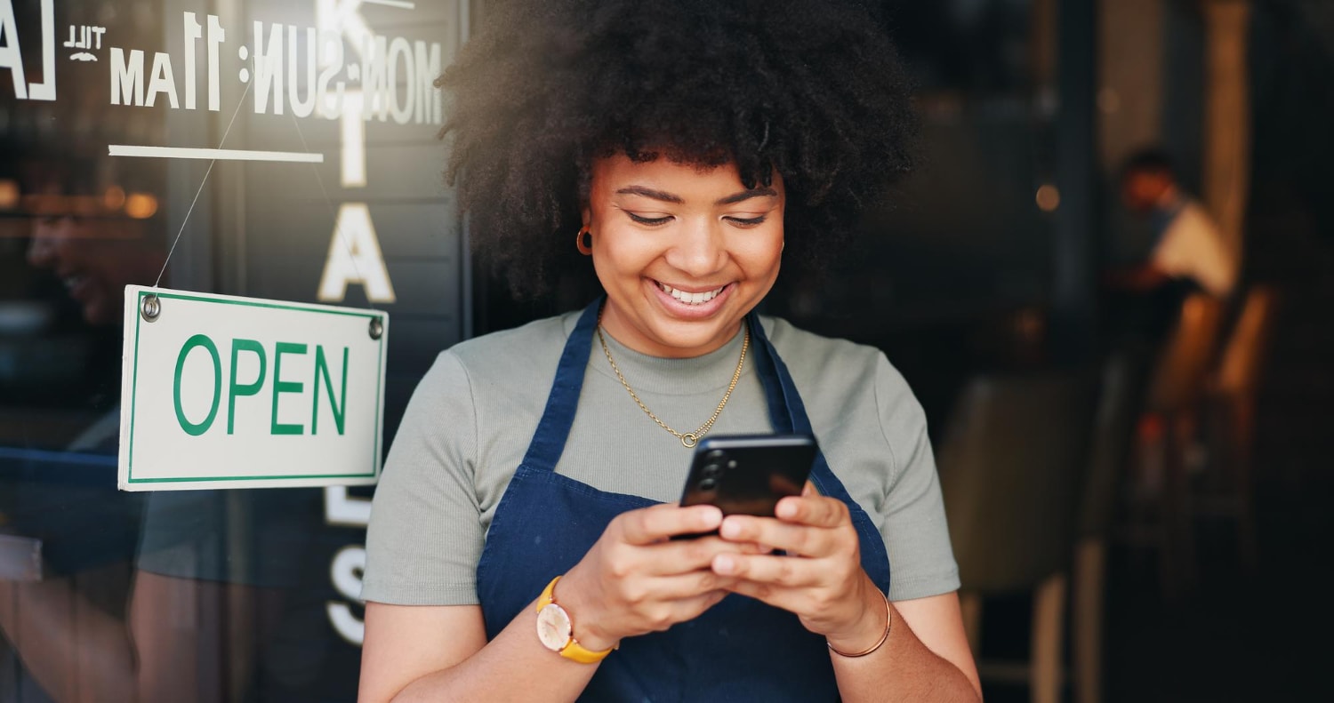 A woman wearing a dark blue apron and green shirt stands outside a storefront with an “Open” sign, smiling at her phone. This moment captures the effectiveness of business texting for customer communication in a retail setting.