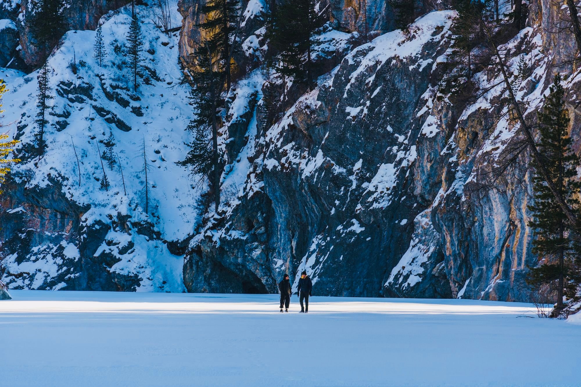 Two hikers in winter boots cross a frozen landscape, surrounded by snow-covered cliffs.