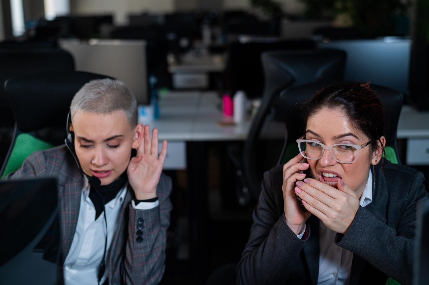 Two call center agents wearing headsets engage in a stressful customer interaction. One woman cups her hand over her mouth while speaking into the phone, while her colleague looks frustrated. This moment highlights the need for real-time contact center analytics to improve service quality.