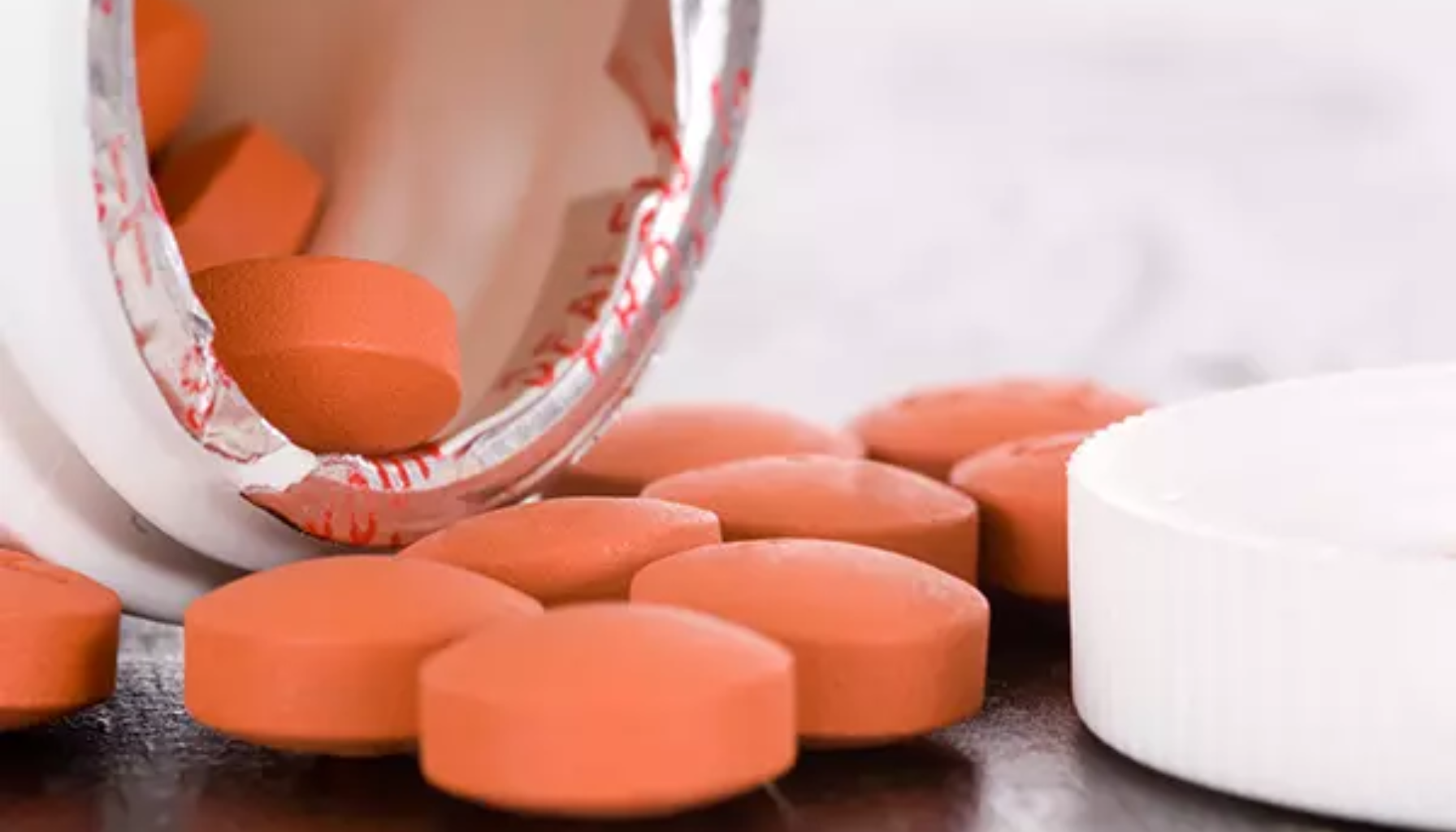 Åpen medisinflaske med røde runde tabletter som ligger spredt på bordet ved siden av hvitt lokk.