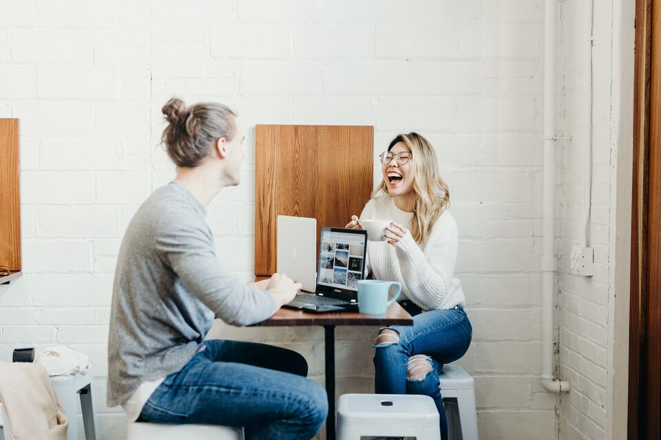 Zijaanzicht van een man en vrouw die aan een smalle werktafel zitten. Er staan laptops op tafel en de vrouw heeft een kopje vast en lacht.
