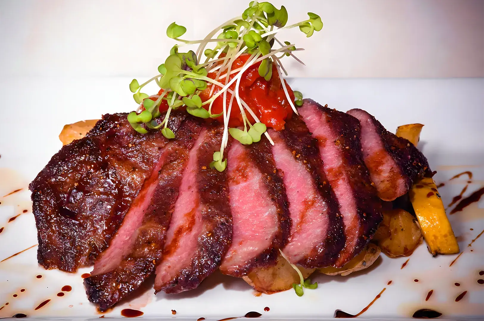 Sliced medium-rare steak on roasted potatoes, garnished with fresh green sprouts and a dollop of red sauce, drizzled with balsamic glaze on a white plate.