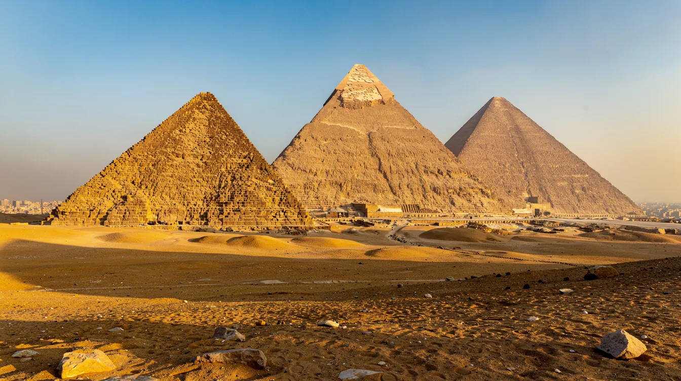 A imagem mostra as majestosas pirâmides de Gizé, localizadas no Egito, com a grande pirâmide de Quéops em destaque, cercada por um deserto árido e um céu azul. Essas estruturas impressionantes, que datam de milhares de anos a.C., são um importante símbolo da história e cultura do mundo antigo.