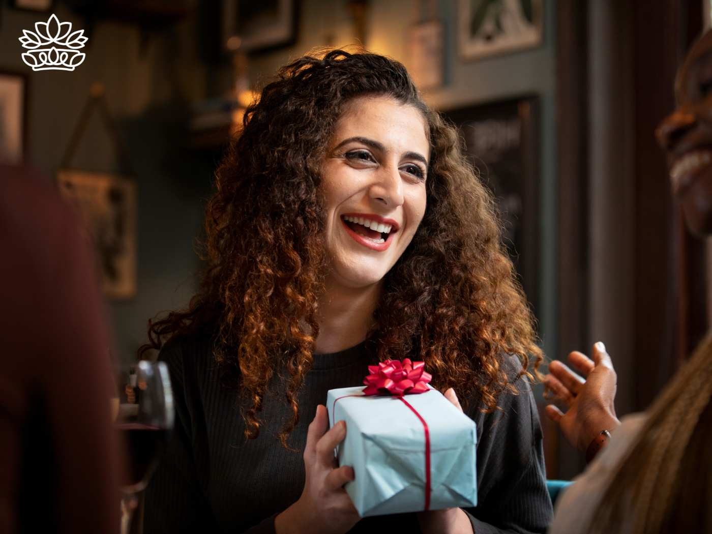 Joyful woman with curly hair laughing and holding a gift box at a social gathering, exemplifying the delight of the Gift Boxes by Recipient Collection from Fabulous Flowers and Gifts.