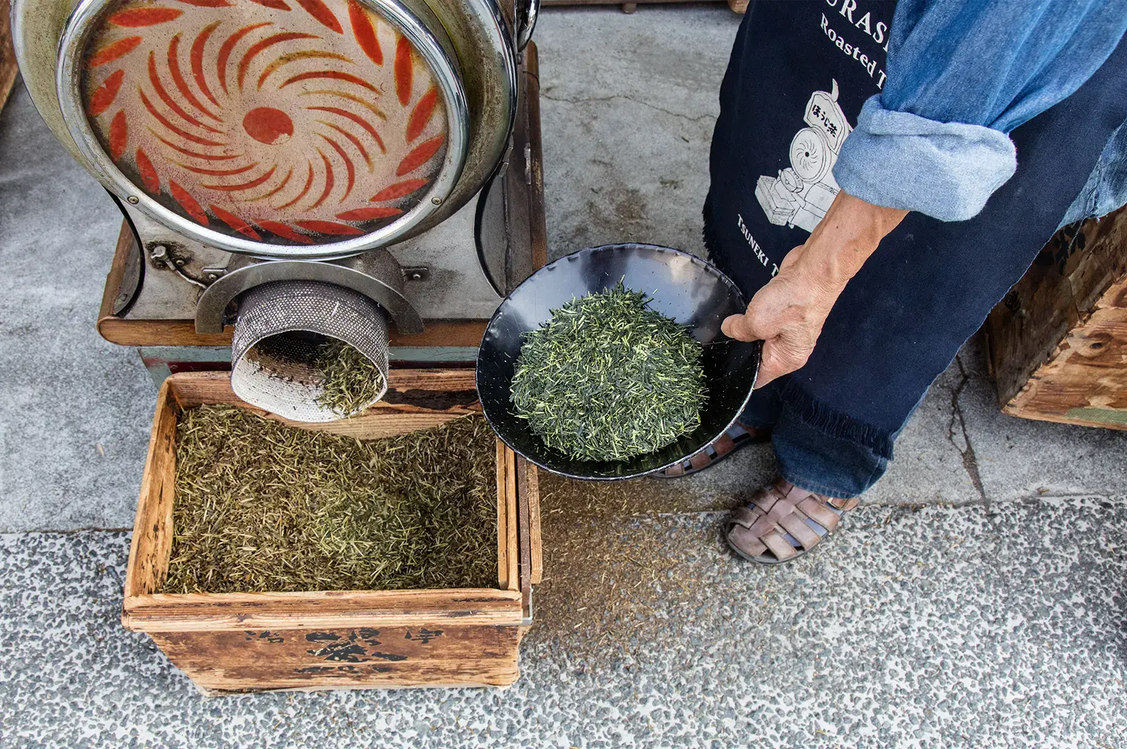 A person wearing a denim apron holds a black bowl filled with deep green tea leaves, positioned next to a large metal tea roasting machine that is currently dispensing roasted, brown tea leaves into a wooden box below.