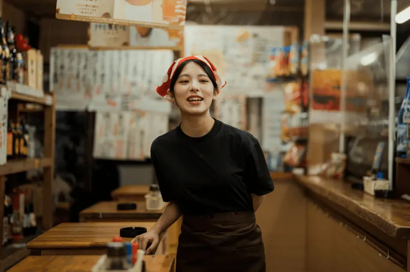 A candid shot of a young female izakaya staff member wearing a red and white patterned bandana, smiling warmly from behind a wooden service counter in a dimly lit, cozy restaurant.