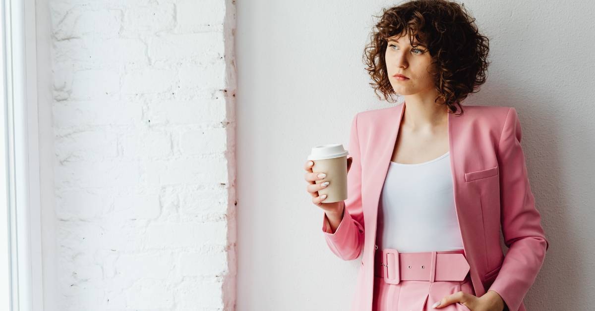 A woman in a pink suit holding a coffee cup, looking thoughtfully out of a window, contemplating her business tax certificate.