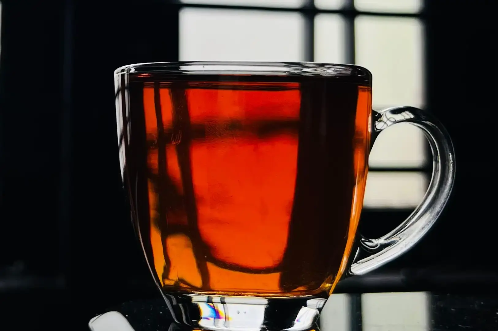 A clear glass mug filled with rich, glowing orange-amber tea sits on a reflective surface against a dark background with window-frame reflections catching the light.