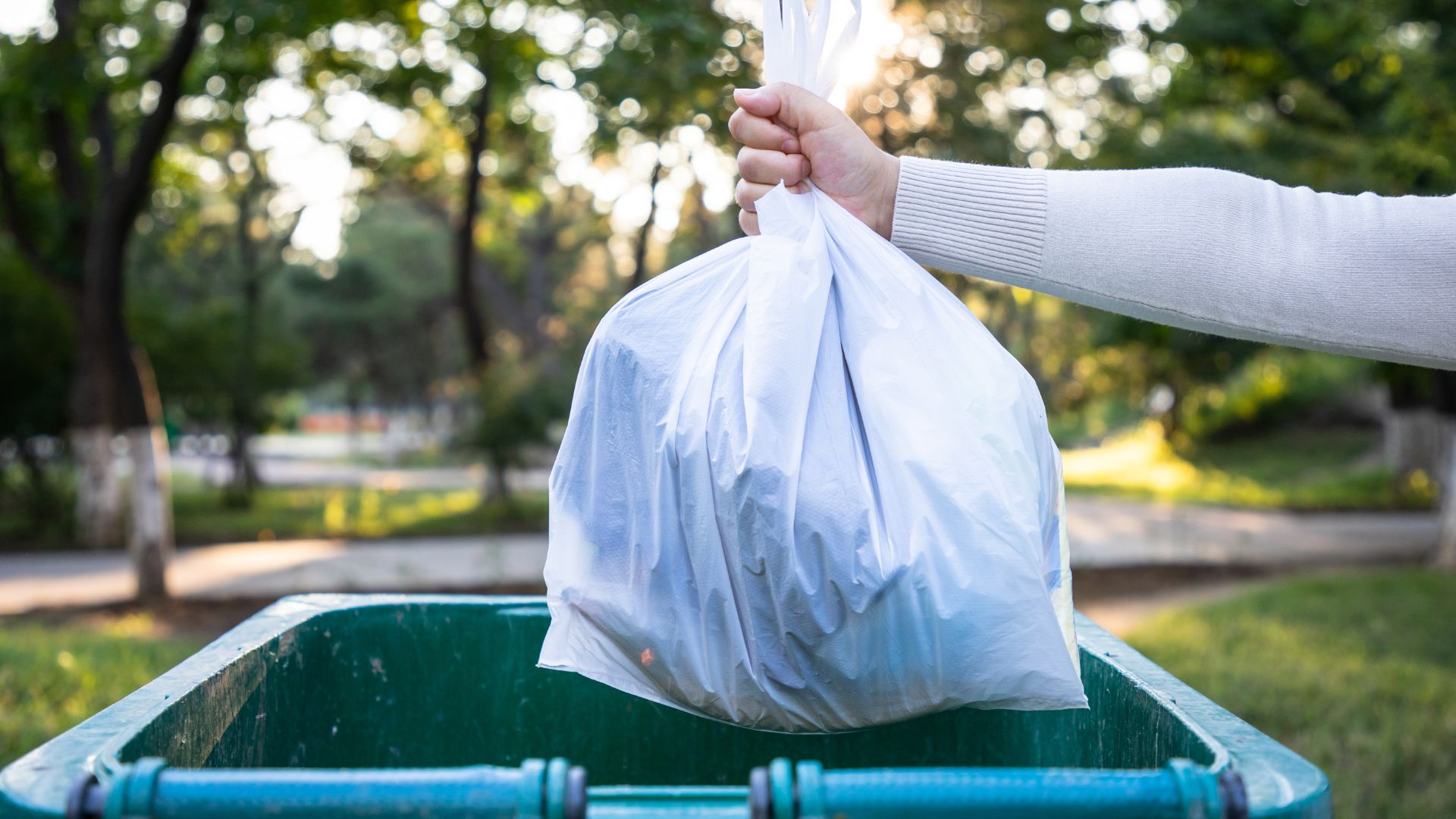 An image of a person throwing away a bag of trash into a garbage container with a lid.