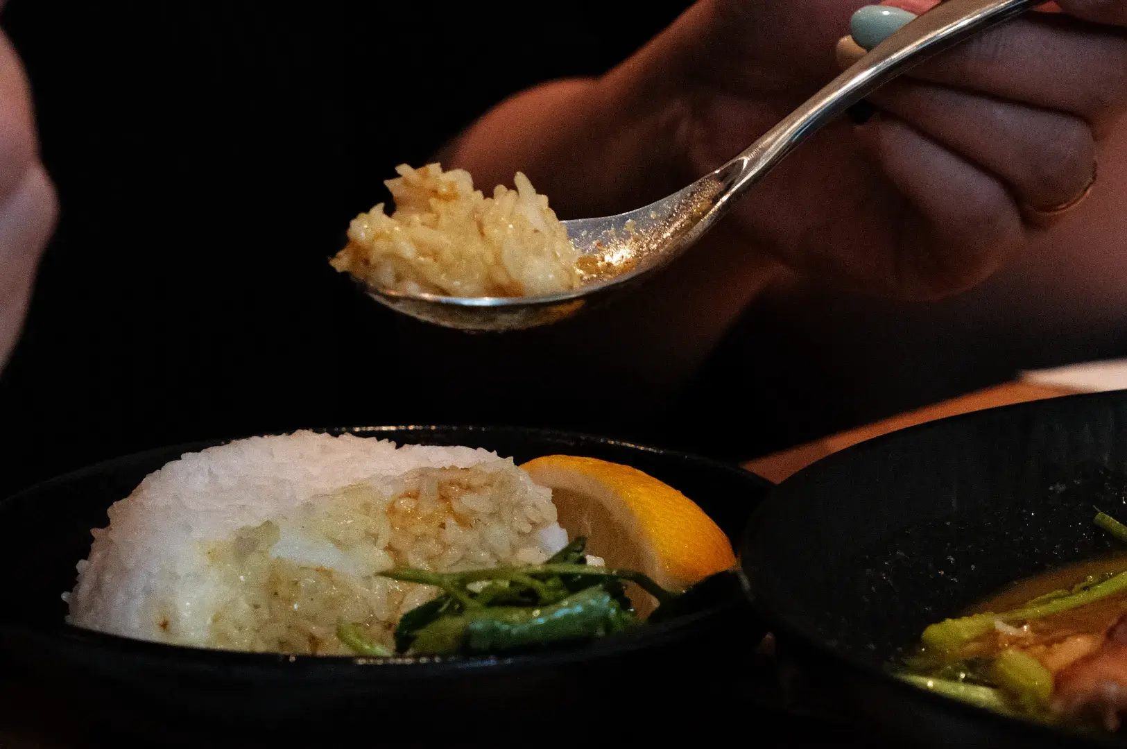 A hand lifts a spoonful of rice above a plate of rice, greens, and lemon beside a bowl of soup, creating a warm, intimate dining scene.