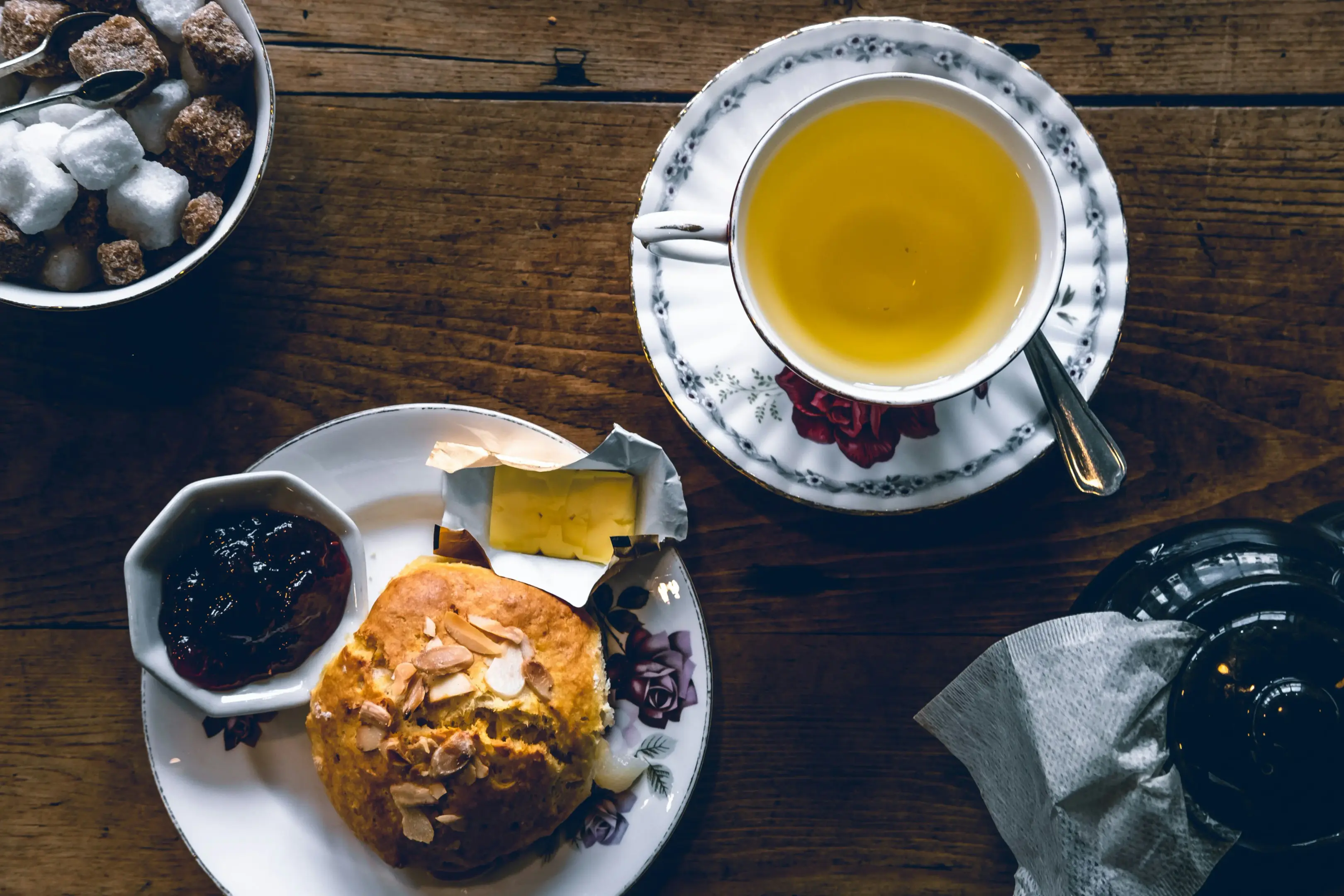 A top-down view shows a rustic wooden table set with a floral teacup of golden tea, a teapot, and a bowl of sugar cubes. Beside the tea sits a plate featuring a sliced scone topped with slivered almonds, accompanied by small servings of jam and butter.