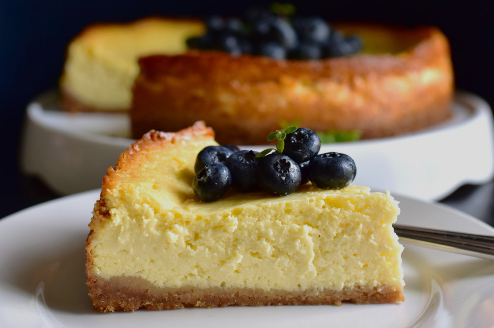 A creamy slice of cheesecake topped with fresh blueberries sits on a white plate, with the rest of the cheesecake in the background, creating a tempting scene.