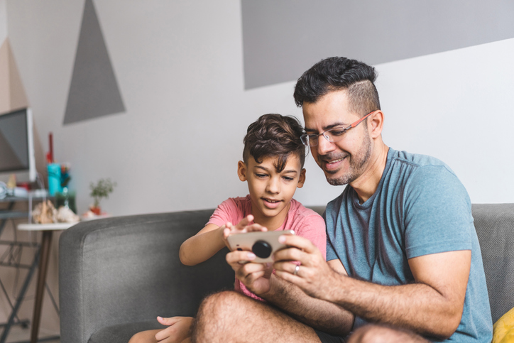 Cheerful young dad and son, both with dark hair, sitting on the sofa watching something on a cell phone. 