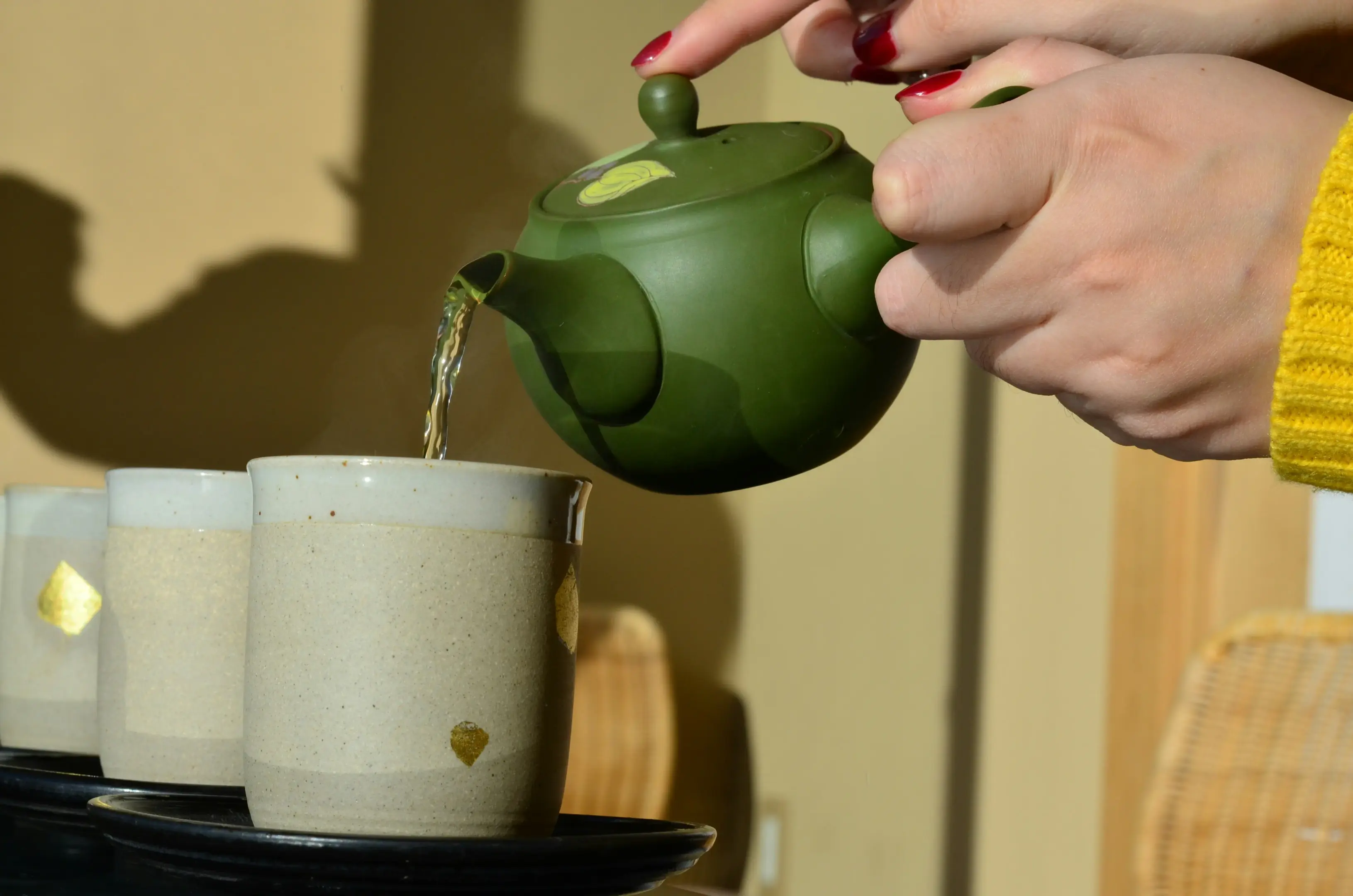 A pair of hands carefully pours steaming tea from a green ceramic teapot into a series of cream-colored cups adorned with gold leaf. The scene is bathed in warm sunlight, casting soft shadows against a neutral background.