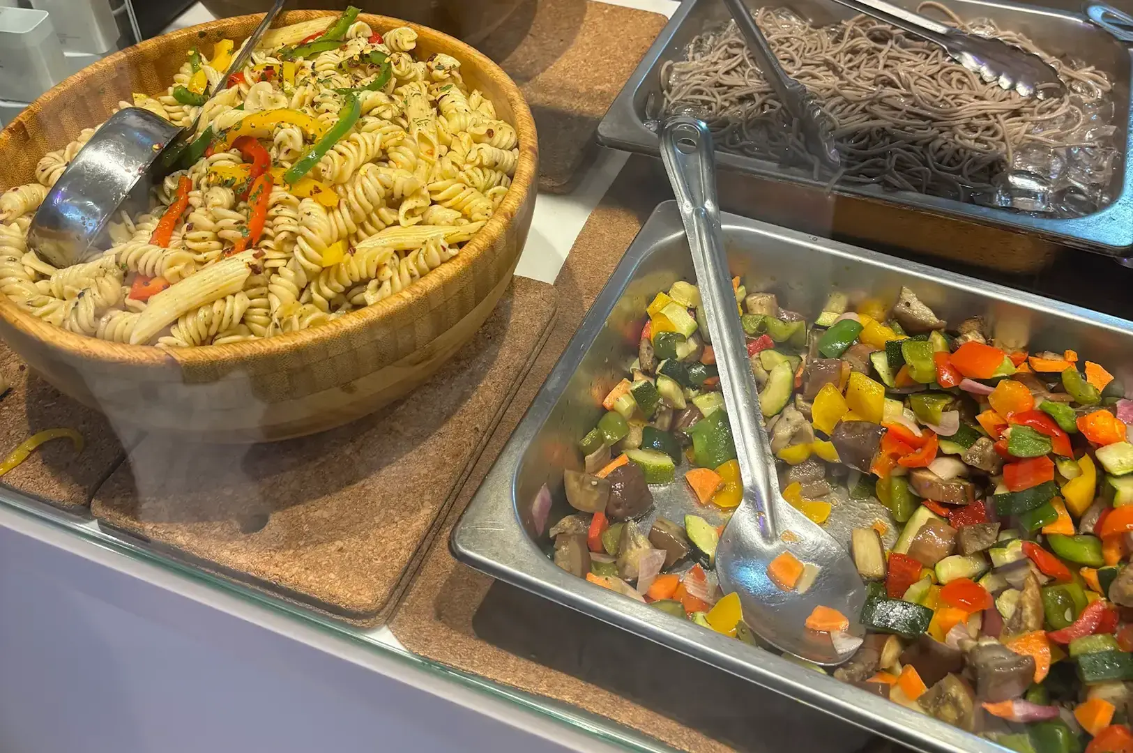 Al Marche food station with wooden bowl of fusilli pasta, grilled vegetables in metal tray, and soba noodles