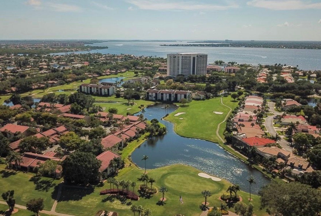 The image depicts luxury waterfront condominiums in Fort Myers, featuring private balconies that offer stunning views of the Intracoastal Waterway, surrounded by lush landscapes and rolling hills. This gated community includes amenities such as a heated competition size pool, tennis club, and waterfront restaurant, catering to various lifestyle needs.