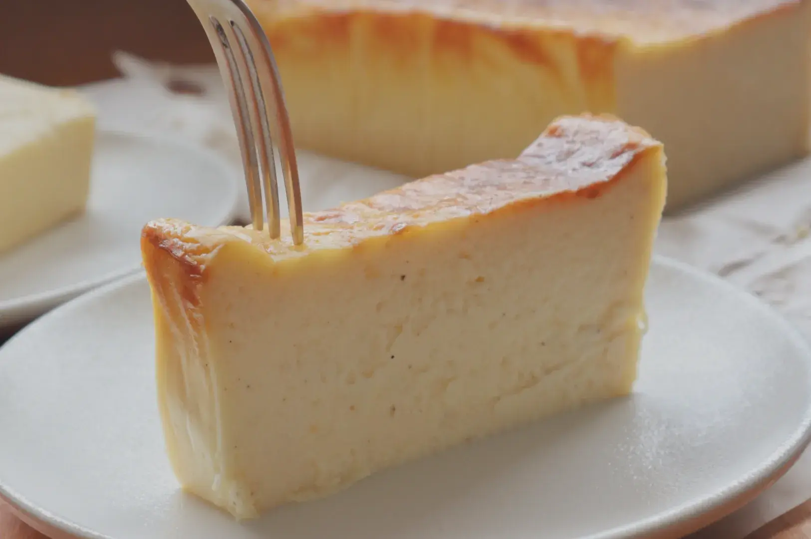 A slice of creamy cheesecake on a white plate with a fork inserted, other slices visible in the background.