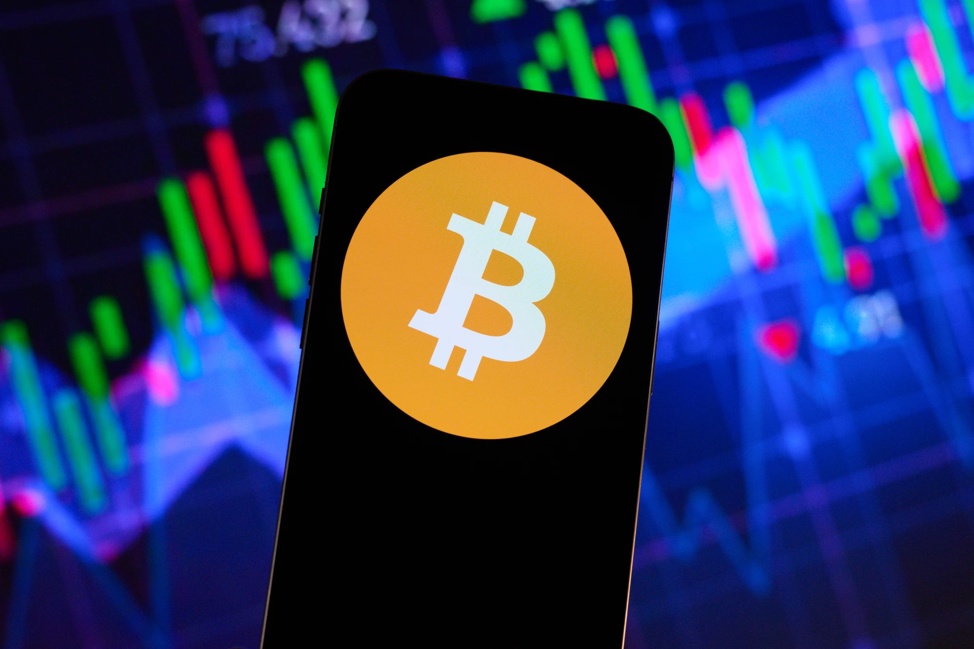 The Bitcoin logo is seen on a silhouette of a smart phone against a background of stock trading graphs.