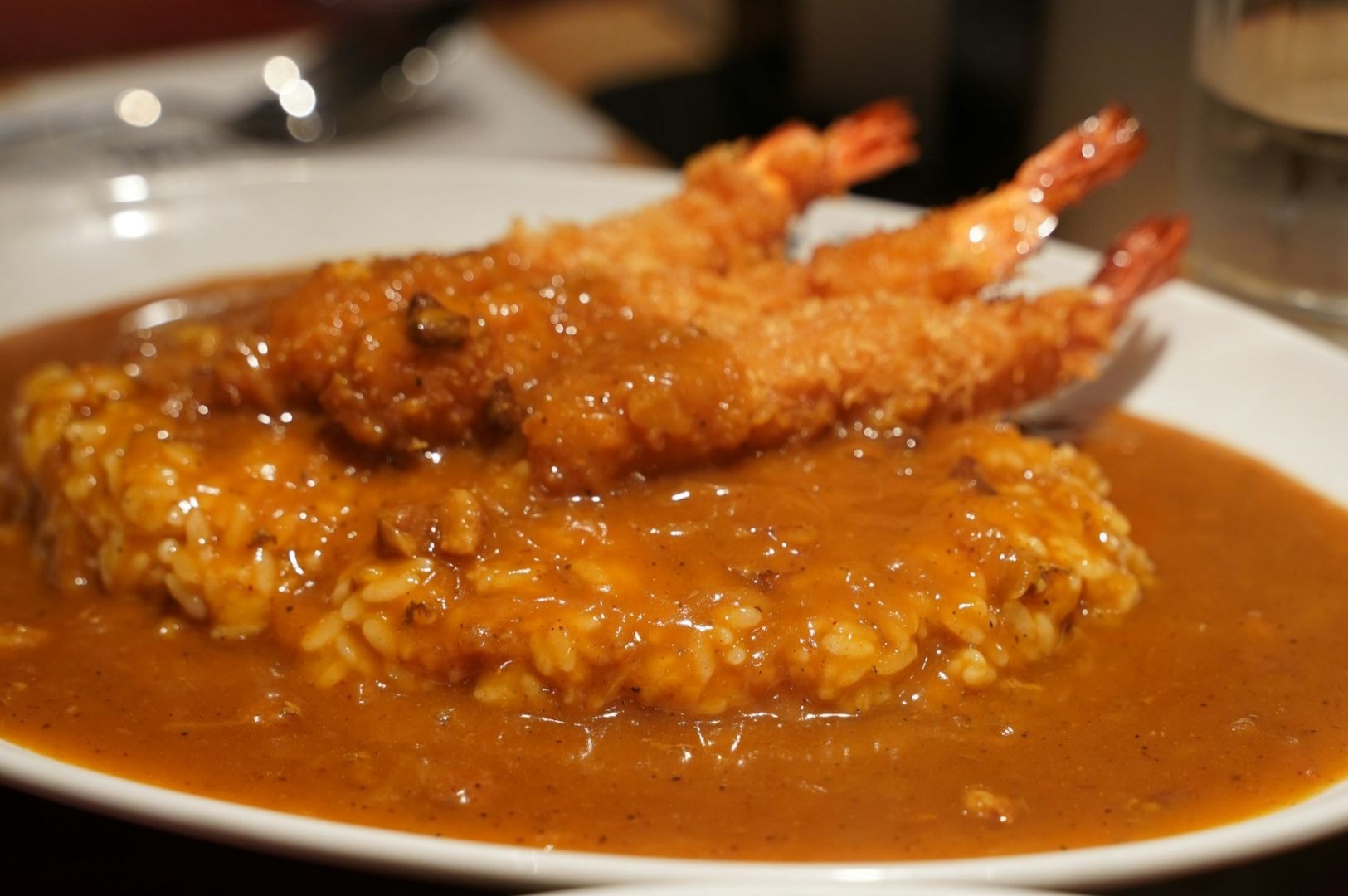 A plate of Japanese curry rice with two golden, crispy fried shrimp on top, covered in rich brown curry sauce, set against a blurred background.