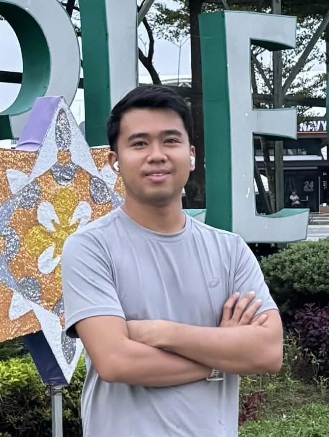 A young man in athletic wear — gray t-shirt, dark shorts, white crew socks, and bright neon green running shoes — stands with arms crossed and a calm smile in front of the large Megaworld Maple Grove landmark sign in the Philippines, surrounded by manicured hedges and decorative star ornaments under an overcast sky.