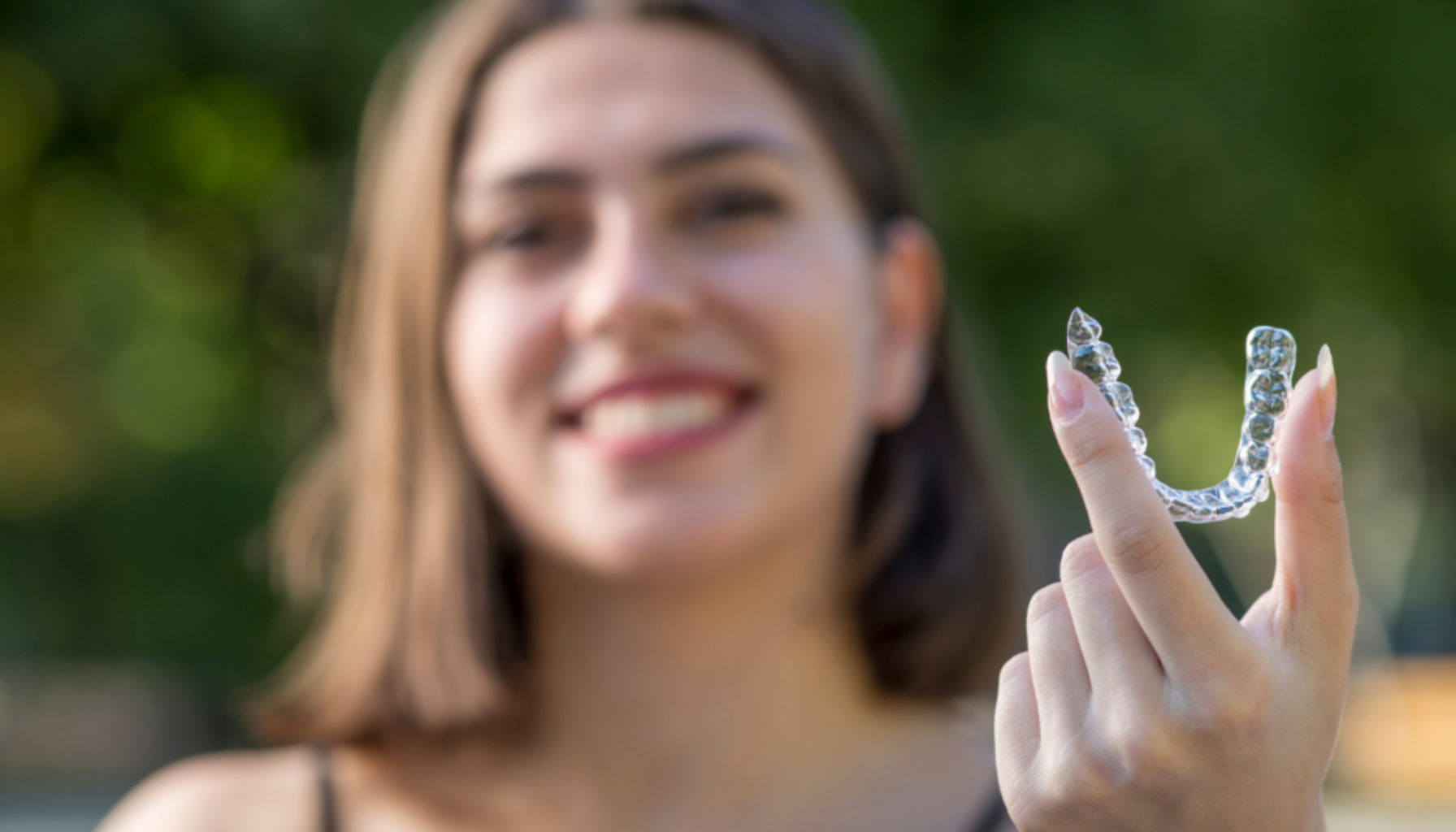 Ung kvinne holder en usynlig tannregulering i hånden og smiler utendørs med grønn bakgrunn.