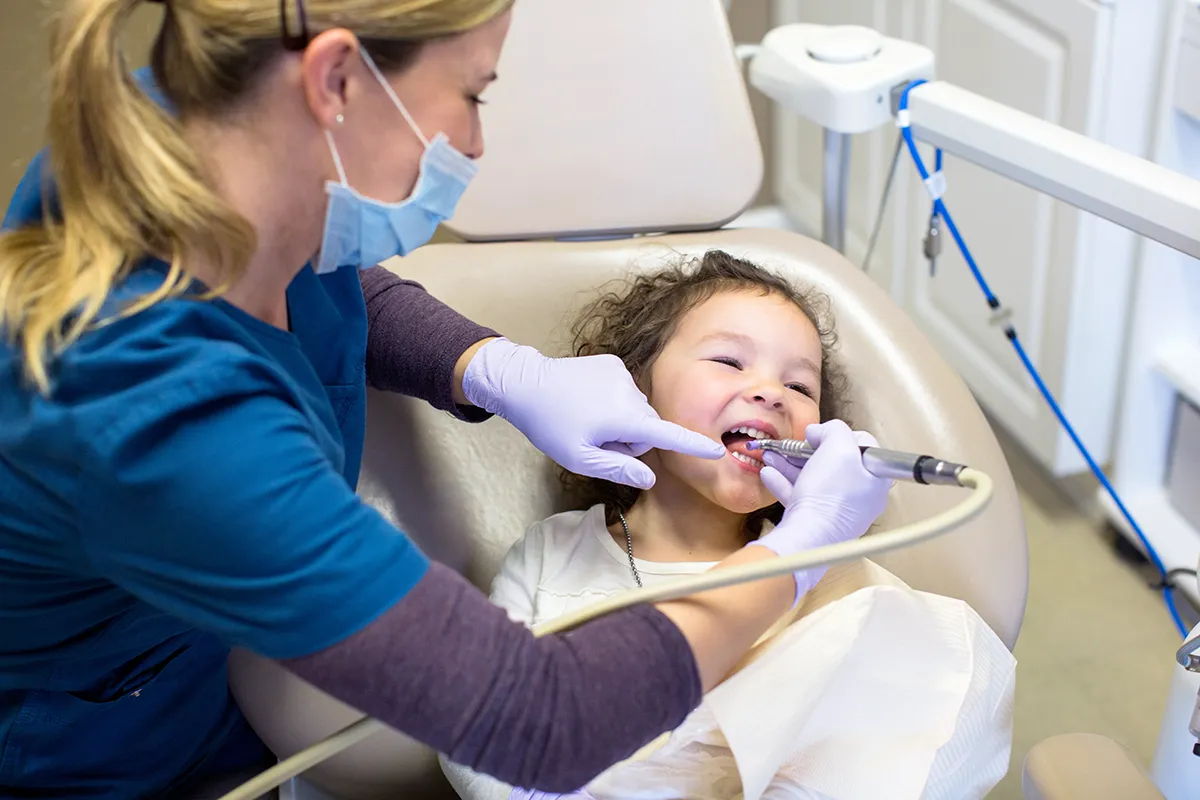 Smiling child during dental cleaning with masked professional in medical setting.