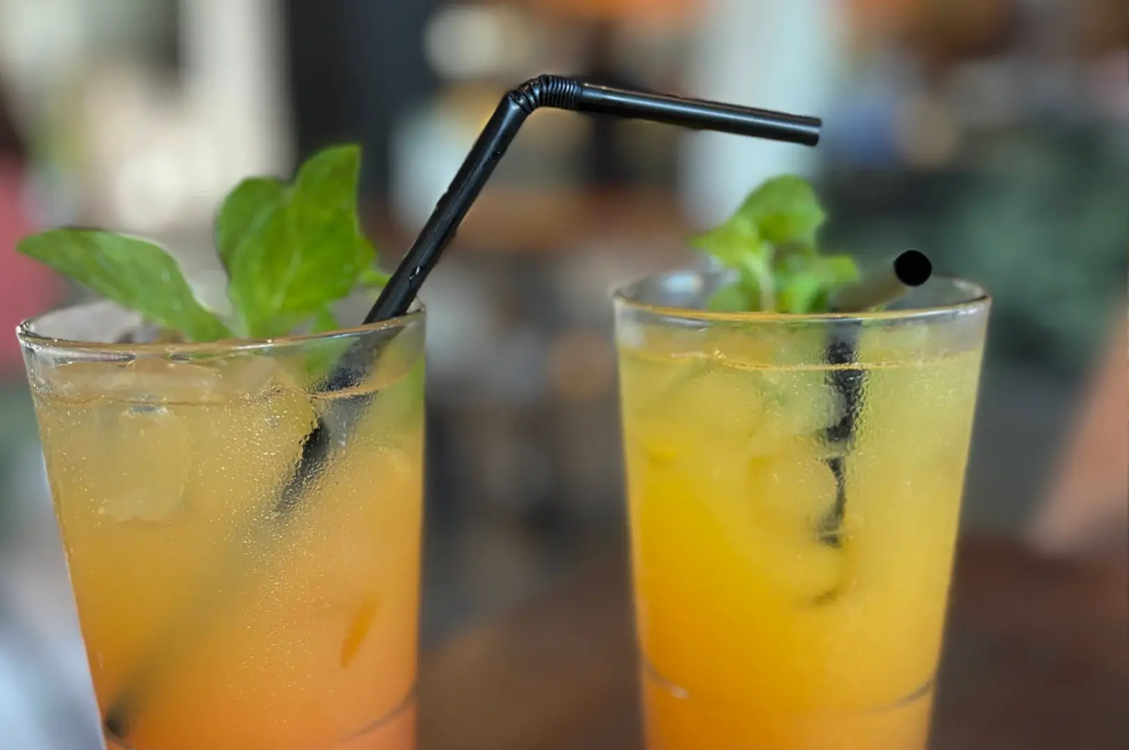 A close-up photograph showcasing two tall, clear glasses filled with vibrant, icy orange-yellow citrus drinks resting on a wooden surface. The glass on the left has a soft gradient transition from a darker orange at the base to a lighter yellow-orange at the top, covered in condensation, with a black straw bending toward the right and garnished with fresh green mint leaves. The glass on the right is filled with a matching bright yellow liquid and crushed ice, condensation pooling on its exterior, containing a straight black straw and a similar green garnish. The background is softly blurred, keeping the focus entirely on the refreshing, colorful beverages.