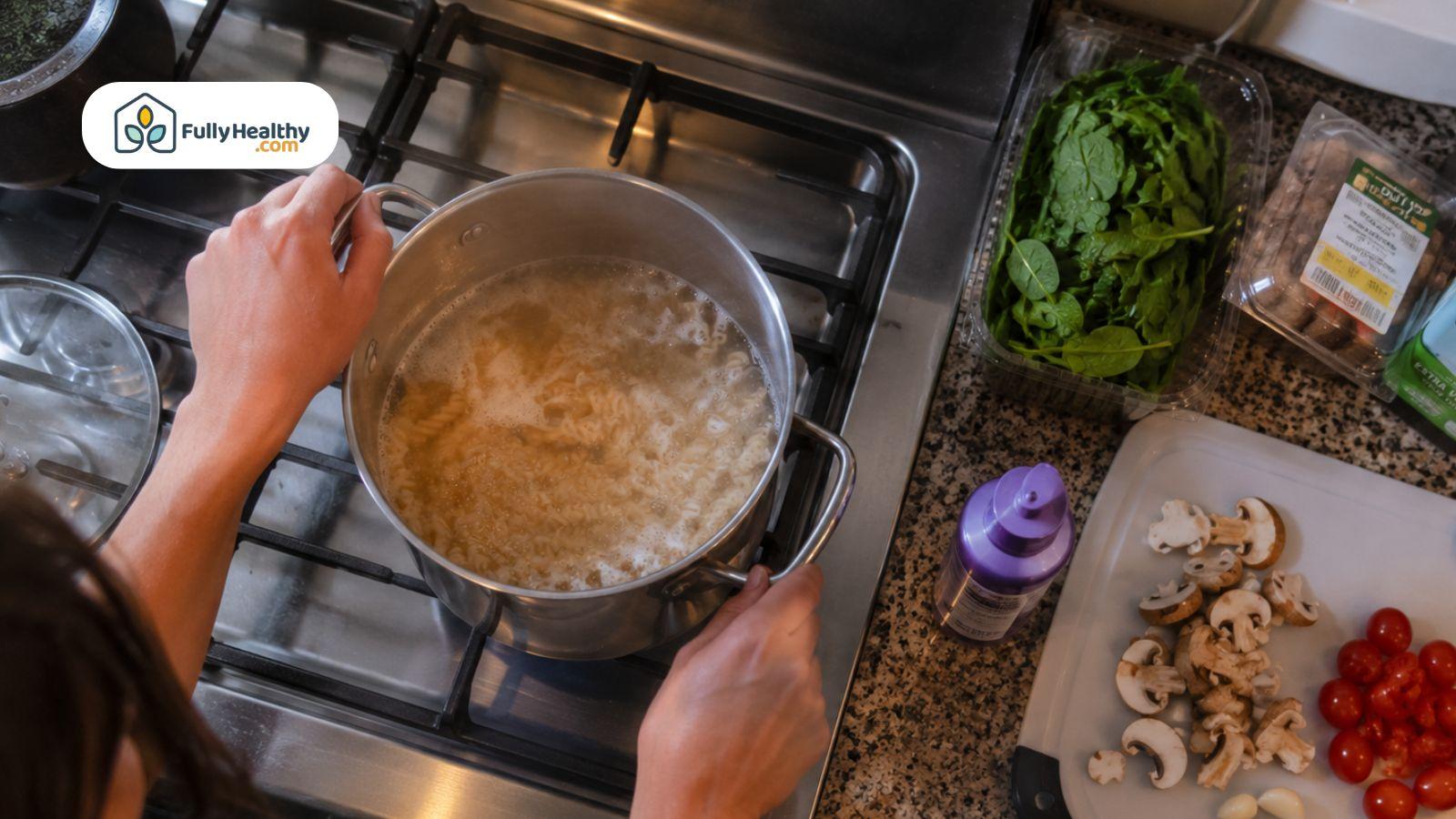 Person cooking pasta on stovetop beside fresh spinach, mushrooms, and cherry tomatoes.