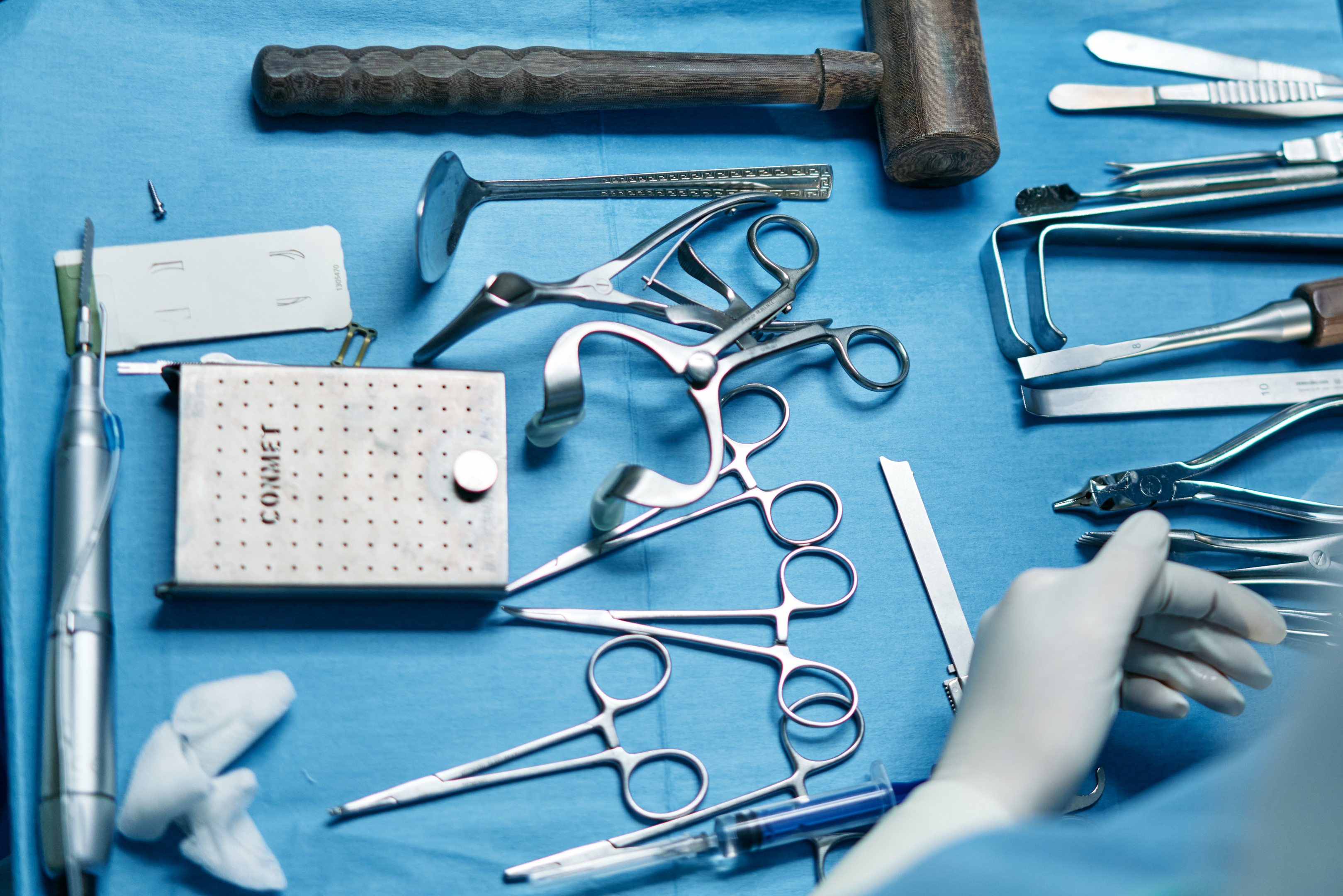 A table covered with a blue cloth, displaying various surgical tools made of surgical stainless steel, with a doctor's gloved hand visible in the bottom right corner.