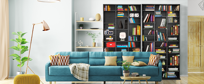 An Artiss Adjustable Black Book Shelf is leaning against the back wall of a living room. It is filled with colourful books and various household items. There is a blue couch and yellow ottoman in the foreground.