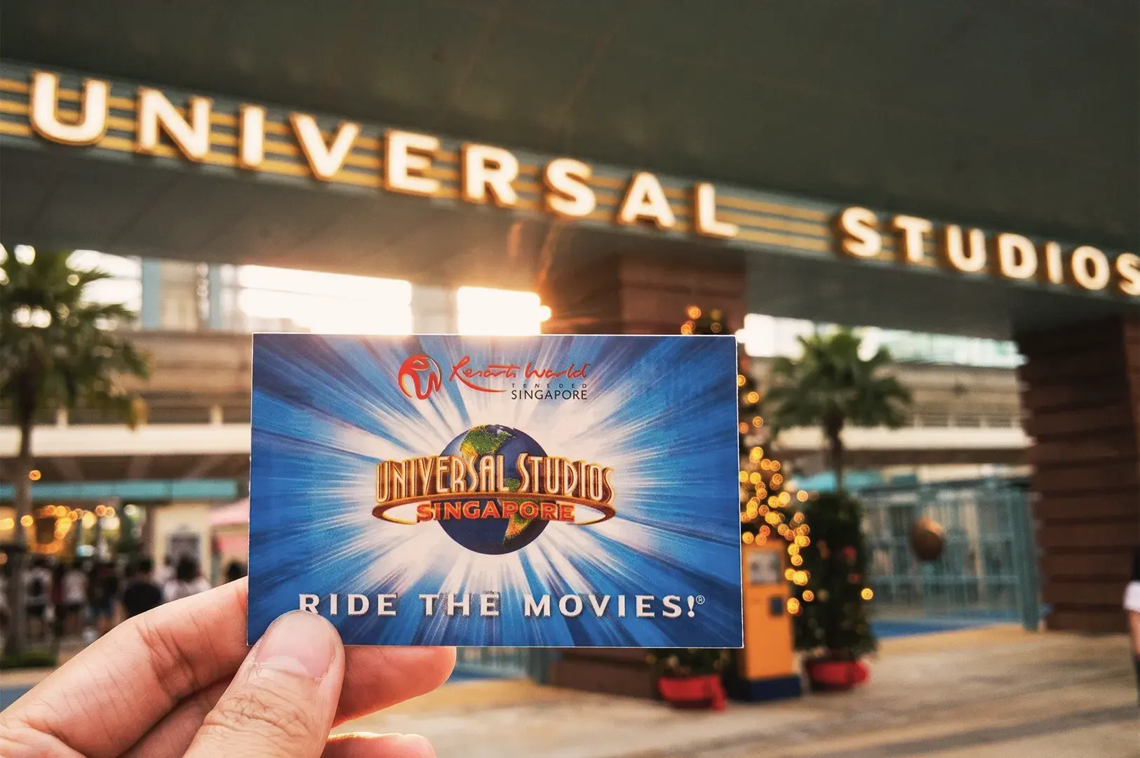 Hand holding Universal Studios Singapore ticket in front of park entrance at Resorts World Sentosa, highlighting “Ride the Movies” slogan.