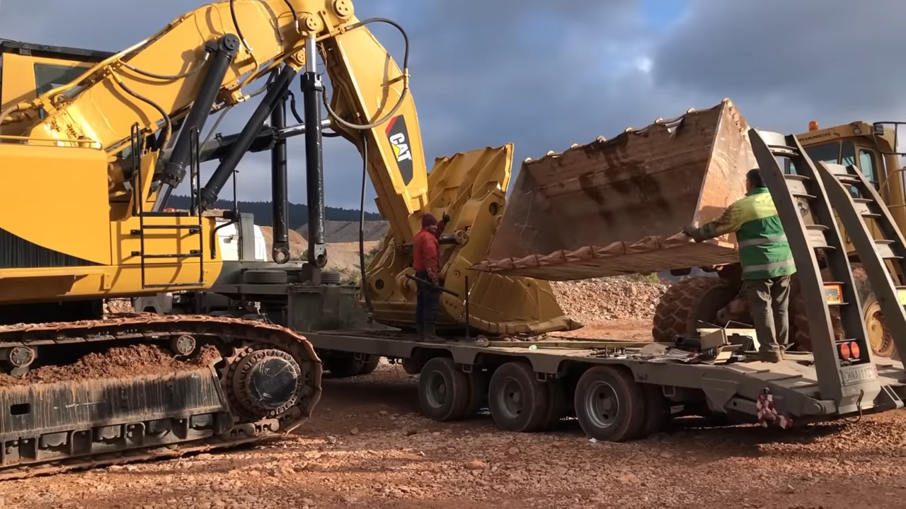 A large yellow excavator with a CAT logo loading a sizable metal scoop bucket onto a flatbed trailer with the assistance of two workers wearing safety vests, one guiding the bucket and the other standing on the trai