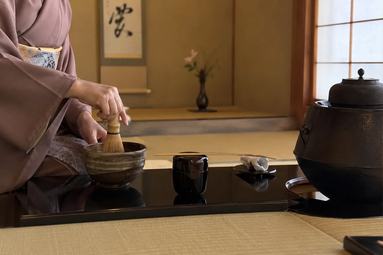 A woman in a muted mauve kimono sits in profile while performing the tea ceremony, her focus entirely on whisking the matcha in a deep bowl. She works on a low, glossy black tray that holds a black tea caddy and a small white cloth neatly folded. The background is simple and uncluttered, with a tall, slender vase of flowers adding a touch of natural elegance to the serene environment.