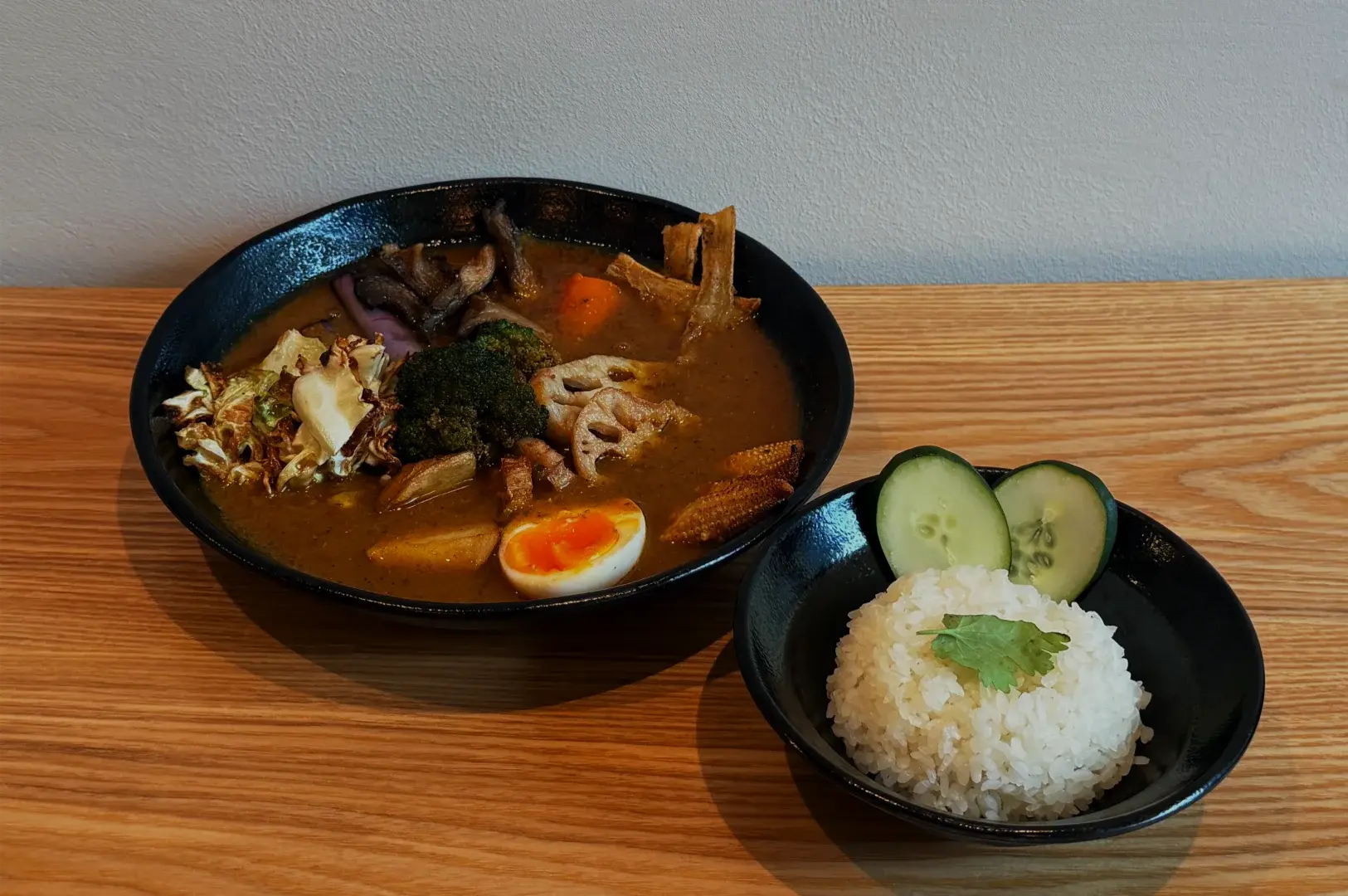 A black bowl of rich vegetable curry with mushrooms, greens, and a soft-boiled egg sits beside rice topped with cilantro and cucumber; a cozy meal.