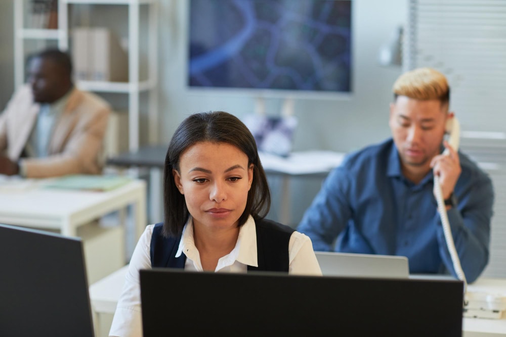 Three office workers concentrate at their desks, with one man using a corded phone and two others focused on their screens in a tech-driven office.
