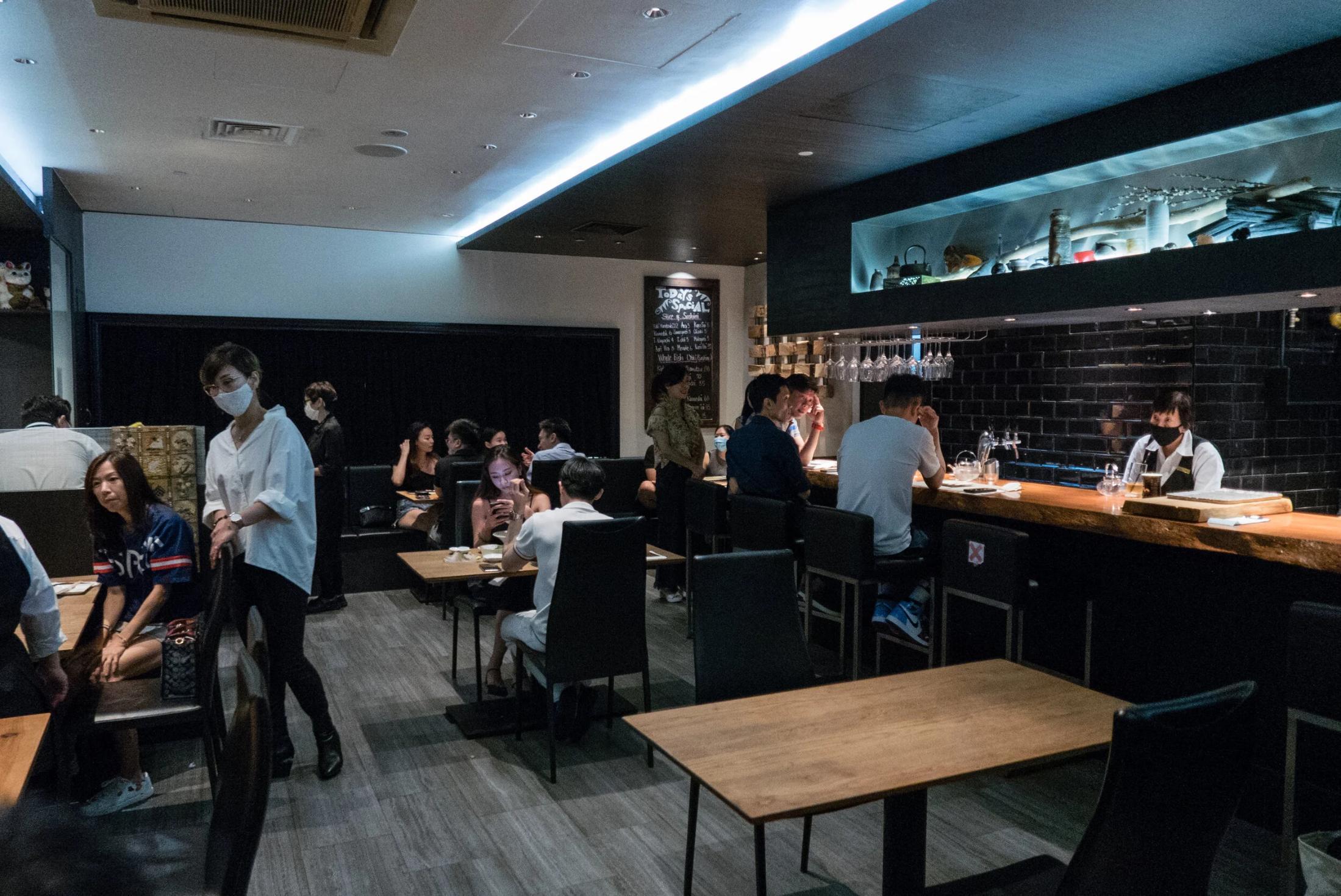 The image captures the bustling interior of a modern, dimly lit restaurant featuring a sleek bar and wooden dining tables. Patrons and staff, some wearing face masks, are seen engaging in a contemporary dining atmosphere with dark decor and recessed lighting.